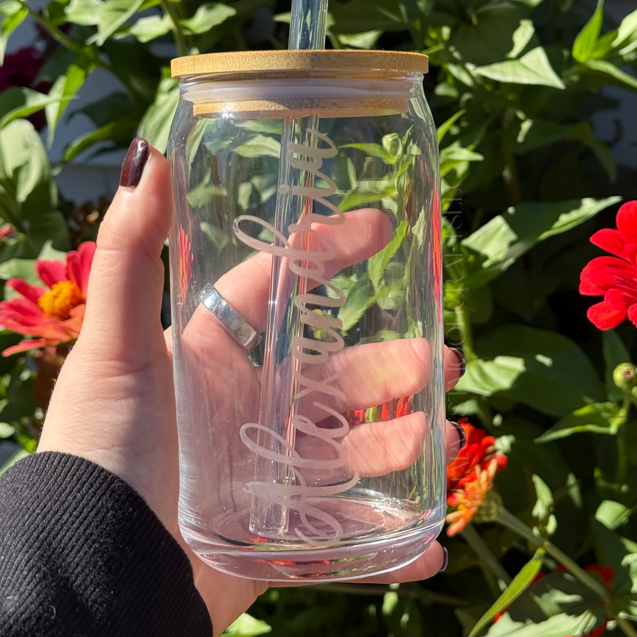 Clear glass tumbler with bamboo lid and straw held by a hand in front of flowers